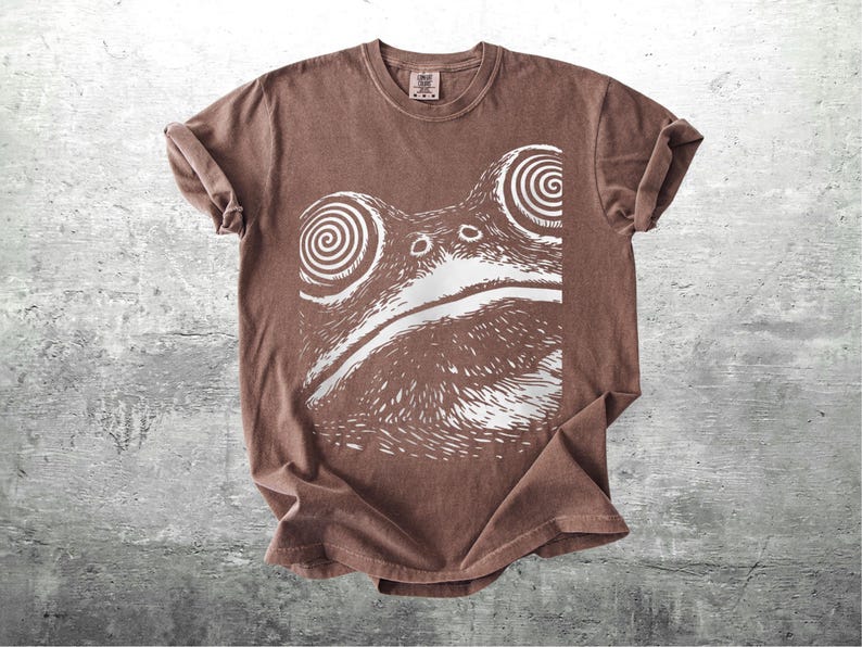 May include: Brown t-shirt featuring a white graphic of a frog's face with hypnotic spiral eyes. The shirt has a crew neck and short sleeves. The fabric appears soft and slightly faded, giving it a vintage look.