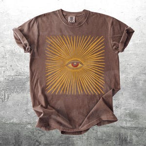May include: Brown t-shirt featuring a yellow sunburst design with a red eye in the center. The shirt has a crew neck and short sleeves. The graphic is centered on the front of the shirt.