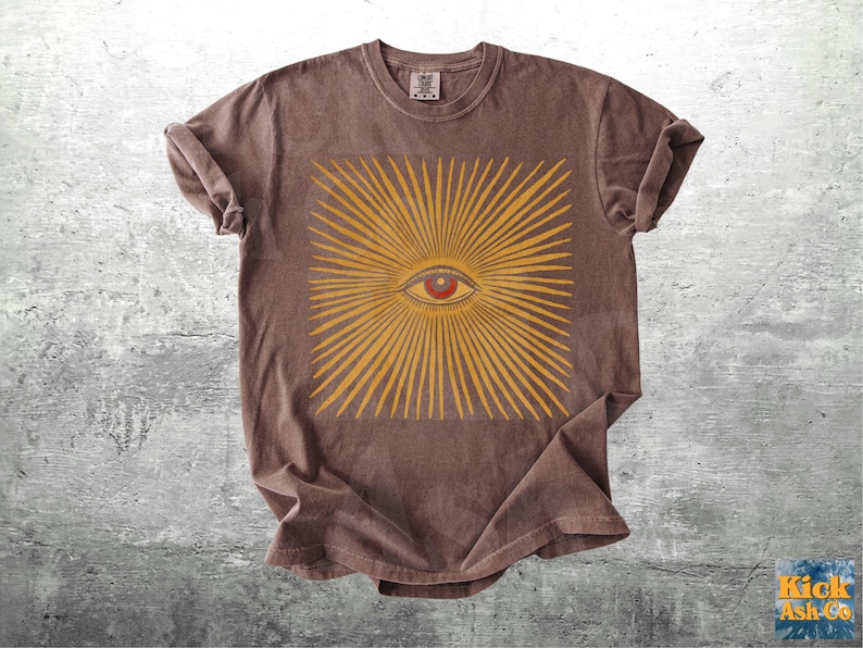 May include: Brown t-shirt featuring a yellow sunburst design with a red eye in the center. The shirt has a crew neck and short sleeves. The graphic is centered on the front of the shirt.