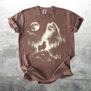 May include: Brown t-shirt featuring a graphic of a Pomeranian dog howling at the moon. The design includes a full moon, trees, and stars, creating a night scene. The shirt has a crew neck and short sleeves.