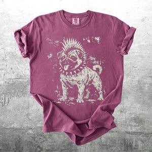 May include: A maroon t-shirt featuring a punk rock pug graphic. The dog has a mohawk, spiked collar, and a tongue sticking out. The graphic is distressed, giving it a vintage look. The shirt is folded on a gray surface.