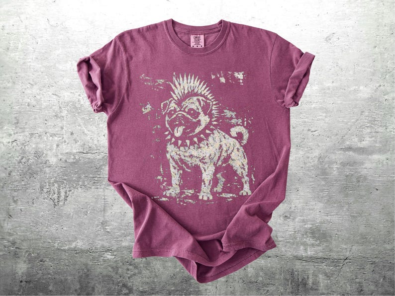 May include: A maroon t-shirt featuring a punk rock pug graphic. The dog has a mohawk, spiked collar, and a tongue sticking out. The graphic is distressed, giving it a vintage look. The shirt is folded on a gray surface.
