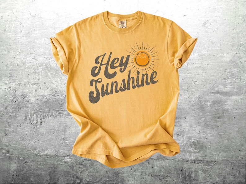 May include: Mustard yellow t-shirt with rolled sleeves. The shirt features the text "Hey Sunshine" in a dark gray, vintage-style font, with a sun graphic. The shirt is laid flat on a gray surface.