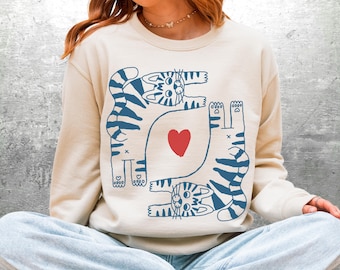 Puede incluir: Sudadera color crema con un diseño de gato azul y un corazón rojo. El gráfico incluye tres gatos en varias poses. Una parte superior casual y cómoda para los amantes de los gatos.