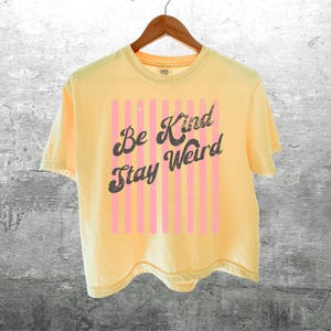 May include: A pale yellow short-sleeve t-shirt featuring a graphic design with vertical light pink stripes and the dark grey text "Be Kind Stay Weird" printed in a cursive font across the centre, hanging on a wooden hanger.