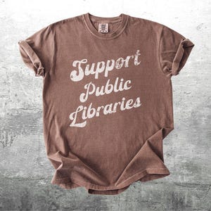 May include: A brown t-shirt with the words "Support Public Libraries" in a distressed white font. The shirt is made of a soft material and has a crew neck. The sleeves are rolled up.