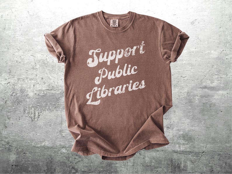 May include: A brown t-shirt with the words "Support Public Libraries" in a distressed white font. The shirt is made of a soft material and has a crew neck. The sleeves are rolled up.