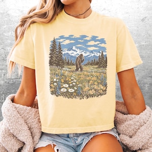 Puede incluir: Camiseta amarillo claro con un gr&aacute;fico de Bigfoot en un prado con monta&ntilde;as y &aacute;rboles. La ilustraci&oacute;n es de estilo vintage, con cielo azul y nubes blancas. La camiseta es de manga corta y cuello redondo.