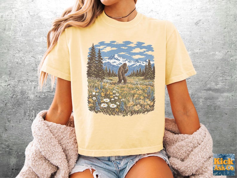 Puede incluir: Camiseta amarillo claro con un gr&aacute;fico de Bigfoot en un prado con monta&ntilde;as y &aacute;rboles. La ilustraci&oacute;n es de estilo vintage, con cielo azul y nubes blancas. La camiseta es de manga corta y cuello redondo.