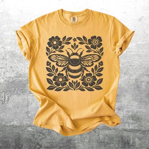 May include: Mustard yellow t-shirt featuring a black and white bee design. The bee is surrounded by floral and leaf elements. The shirt has a crew neck and short sleeves. The fabric appears soft and comfortable.