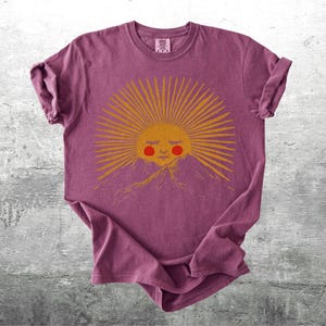 K&ouml;nnte beinhalten: Burgunderfarbenes T-Shirt mit einem goldenen Sonnengrafik, das ein l&auml;chelndes Gesicht und rosige Wangen &uuml;ber einer Bergkette zeigt. Die Sonne hat Strahlen. Das T-Shirt ist gefaltet und hat hochgekrempelte &Auml;rmel.