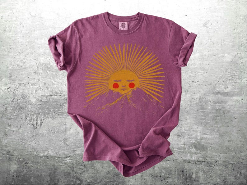K&ouml;nnte beinhalten: Burgunderfarbenes T-Shirt mit einem goldenen Sonnengrafik, das ein l&auml;chelndes Gesicht und rosige Wangen &uuml;ber einer Bergkette zeigt. Die Sonne hat Strahlen. Das T-Shirt ist gefaltet und hat hochgekrempelte &Auml;rmel.
