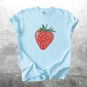 May include: A light blue t-shirt featuring a large, distressed red strawberry graphic with green leaves. The strawberry has white speckles. The shirt is made of soft material and is suitable for casual wear.