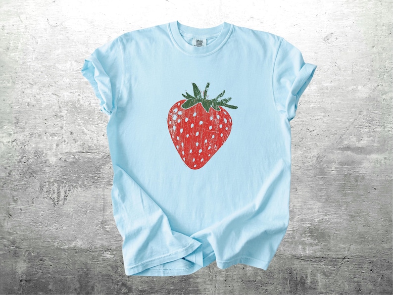 May include: A light blue t-shirt featuring a large, distressed red strawberry graphic with green leaves. The strawberry has white speckles. The shirt is made of soft material and is suitable for casual wear.