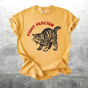 K&ouml;nnte beinhalten: Senfgelbes T-Shirt mit dem Text "FIGHT FASCISM" in einem roten Bogen &uuml;ber einer schwarz-braun gestreiften Katzenzeichnung. Die Katze befindet sich in einer aggressiven Haltung mit offenem Maul. Das Shirt hat hochgekrempelte &Auml;rmel.