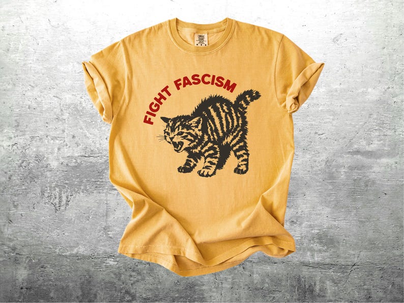 K&ouml;nnte beinhalten: Senfgelbes T-Shirt mit dem Text "FIGHT FASCISM" in einem roten Bogen &uuml;ber einer schwarz-braun gestreiften Katzenzeichnung. Die Katze befindet sich in einer aggressiven Haltung mit offenem Maul. Das Shirt hat hochgekrempelte &Auml;rmel.