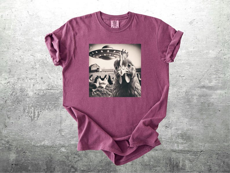 May include: A maroon t-shirt featuring a black and white graphic of a chicken looking at the viewer with a UFO hovering above. Other chickens and a barn are in the background. The shirt is made of soft material.