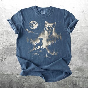 May include: Blue t-shirt featuring a graphic of a chihuahua dog under a full moon and starry night sky. The design includes a silhouette of a dog on a hill and a detailed portrait of a chihuahua. The shirt has rolled sleeves.