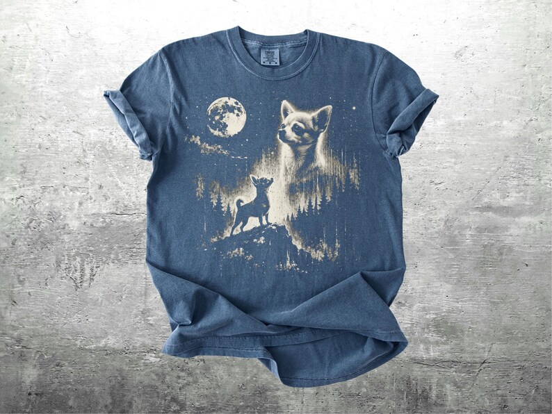 May include: Blue t-shirt featuring a graphic of a chihuahua dog under a full moon and starry night sky. The design includes a silhouette of a dog on a hill and a detailed portrait of a chihuahua. The shirt has rolled sleeves.