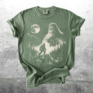 May include: A sage green short-sleeve t-shirt featuring a cream-colored graphic print of a large Bigfoot silhouette, a smaller Bigfoot figure walking through a pine forest, and a full moon set against a starry night sky background.