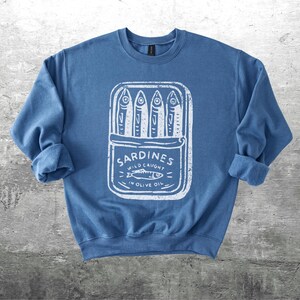 May include: A blue sweatshirt with a white graphic of a sardine can. The can is open, displaying several fish. Text on the can reads "Sardines Wild Caught in Olive Oil". The sleeves are rolled up.