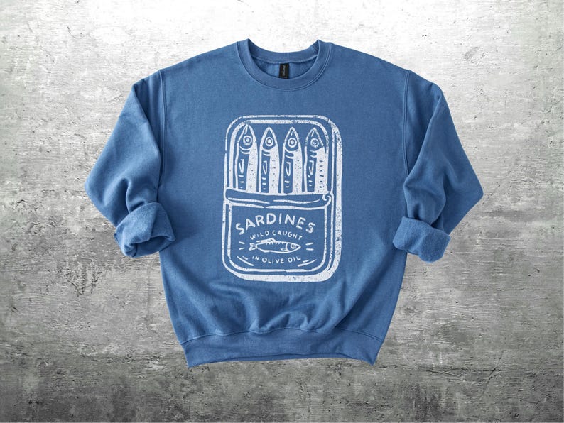 May include: A blue sweatshirt with a white graphic of a sardine can. The can is open, displaying several fish. Text on the can reads "Sardines Wild Caught in Olive Oil". The sleeves are rolled up.