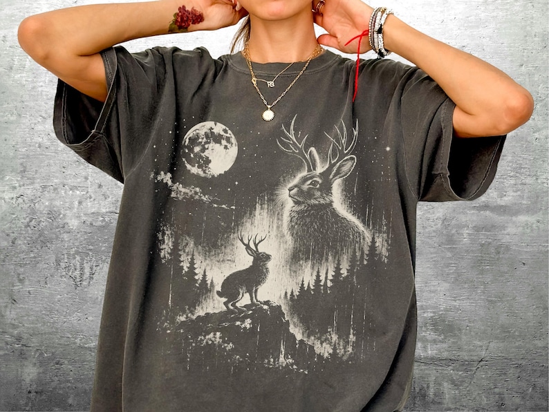 May include: Dark gray t-shirt featuring a detailed graphic of a jackalope with antlers, a full moon, and a forest scene. The design is in white, creating a striking contrast against the shirt's color. The shirt has a relaxed fit.