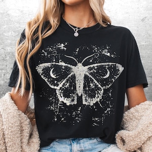 May include: Black t-shirt featuring a distressed white moth graphic with crescent moon details. The design has a vintage, celestial aesthetic. The shirt is part of a clothing collection.