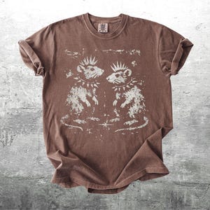 K&ouml;nnte beinhalten: Braunes T-Shirt mit einem verblassten Aufdruck von zwei Punk-Ratten, jede mit Irokesenschnitt und Nietenhalsband. Die Ratten blicken sich an. Das T-Shirt hat einen Rundhalsausschnitt und kurze &Auml;rmel.