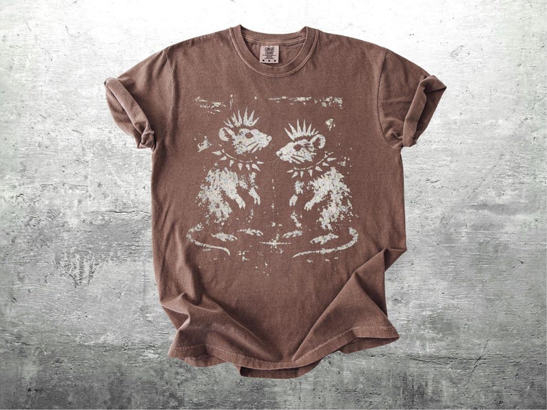 K&ouml;nnte beinhalten: Braunes T-Shirt mit einem verblassten Aufdruck von zwei Punk-Ratten, jede mit Irokesenschnitt und Nietenhalsband. Die Ratten blicken sich an. Das T-Shirt hat einen Rundhalsausschnitt und kurze &Auml;rmel.