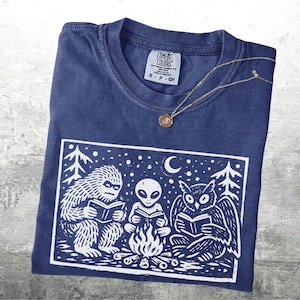 May include: A navy blue Comfort Colors t-shirt with a white graphic of a Bigfoot, an alien, and an owl reading books around a campfire. A gold pendant necklace rests on the shirt.
