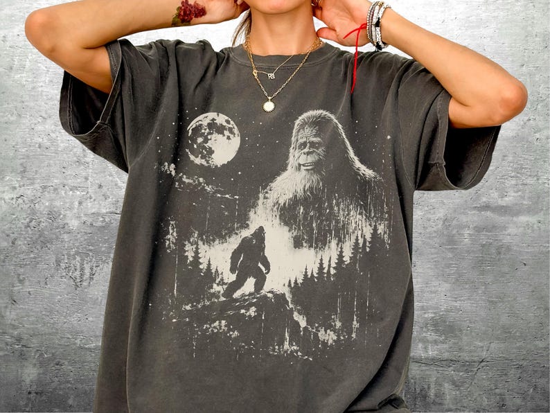 May include: A charcoal gray short-sleeve t-shirt featuring a graphic print of a large Bigfoot creature, a smaller Bigfoot silhouette walking on a rocky hill, a full moon, and a forest of pine trees against a starry night sky background.