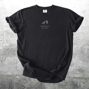 May include: Black t-shirt with rolled sleeves, featuring a small graphic of two birds and the text "attempted murder" in a simple font. The shirt is displayed on a textured gray surface.