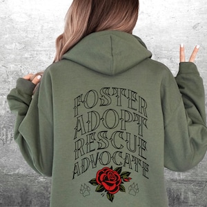 Pu&ograve; includere: Felpa con cappuccio verde con il testo "FOSTER ADOPT RESCUE ADVOCATE" in lettere nere sul retro. Un rosa rossa con foglie verdi e due impronte di zampa nere sono sotto il testo.