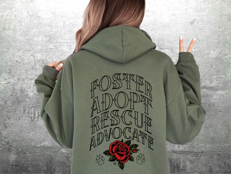 Pu&ograve; includere: Felpa con cappuccio verde con il testo "FOSTER ADOPT RESCUE ADVOCATE" in lettere nere sul retro. Un rosa rossa con foglie verdi e due impronte di zampa nere sono sotto il testo.