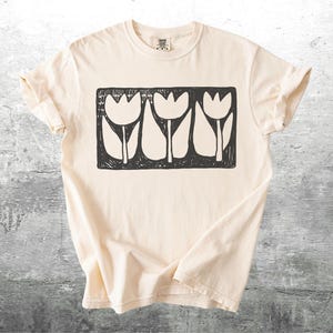 May include: Cream-colored t-shirt featuring a black and white graphic of three stylized tulips within a rectangular frame. The shirt has short sleeves and a crew neck. The fabric appears soft and comfortable.