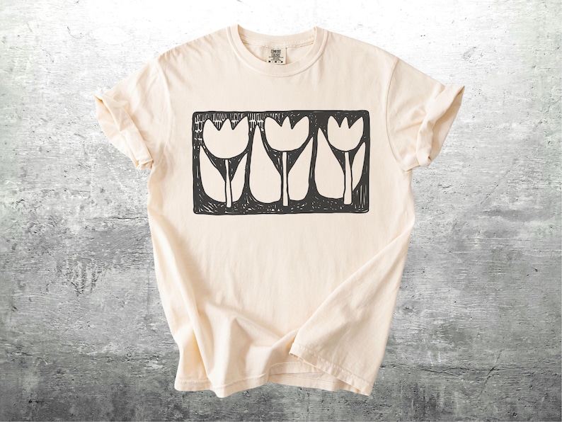 May include: Cream-colored t-shirt featuring a black and white graphic of three stylized tulips within a rectangular frame. The shirt has short sleeves and a crew neck. The fabric appears soft and comfortable.