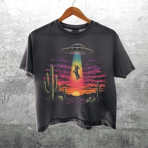 Puede incluir: Una camiseta gris carb&oacute;n con un estampado gr&aacute;fico de un perro salchicha siendo abducido por un OVNI en un paisaje des&eacute;rtico al atardecer. El dise&ntilde;o incluye cactus, un cielo vibrante de color p&uacute;rpura y naranja, y un rayo tractor azul brillante. El texto Kick Ash Co es visible en la esquina inferior derecha.