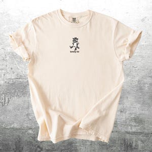 May include: Cream-colored t-shirt with a small black graphic of a cartoon character and the text "howdy-do" underneath. The shirt has a crew neck and short sleeves, and is made of a soft, comfortable material.