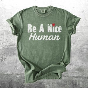 K&ouml;nnte beinhalten: Olivgr&uuml;nes T-Shirt mit dem Schriftzug "Be A Nice Human" in wei&szlig;er, verwaschener Schrift. Ein kleines rotes Herz befindet sich &uuml;ber dem Wort "Nice". Das T-Shirt ist gefaltet, die &Auml;rmel sind hochgekrempelt.