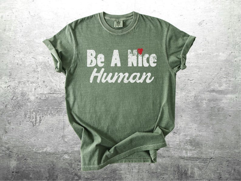 K&ouml;nnte beinhalten: Olivgr&uuml;nes T-Shirt mit dem Schriftzug "Be A Nice Human" in wei&szlig;er, verwaschener Schrift. Ein kleines rotes Herz befindet sich &uuml;ber dem Wort "Nice". Das T-Shirt ist gefaltet, die &Auml;rmel sind hochgekrempelt.