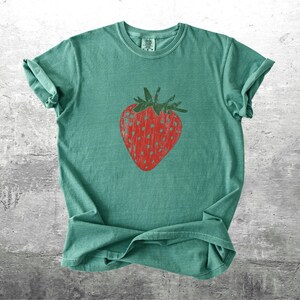 May include: A teal-colored t-shirt featuring a large, red strawberry graphic. The strawberry has a textured appearance with a green stem and leaves. The shirt is made of soft material and has rolled-up sleeves.