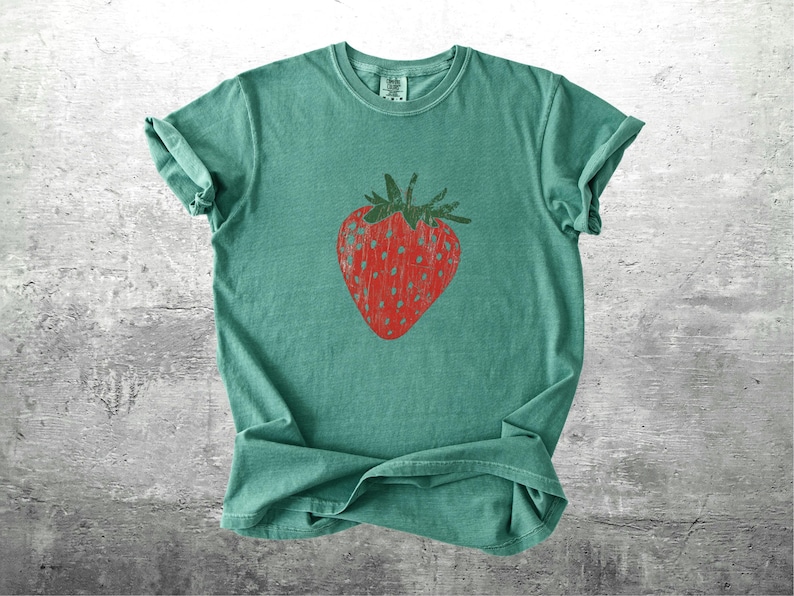 May include: A teal-colored t-shirt featuring a large, red strawberry graphic. The strawberry has a textured appearance with a green stem and leaves. The shirt is made of soft material and has rolled-up sleeves.
