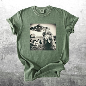 May include: Olive green t-shirt featuring a black and white image of a chicken looking at a UFO. Other chickens and a barn are in the background. The t-shirt has a crew neck and short sleeves.