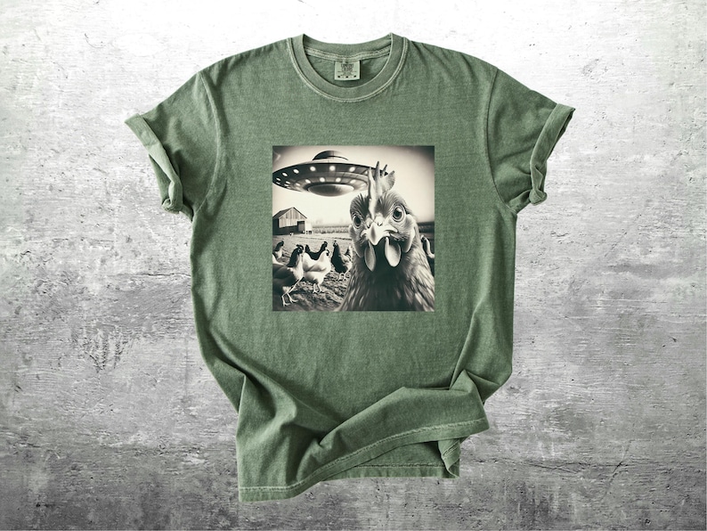 May include: Olive green t-shirt featuring a black and white image of a chicken looking at a UFO. Other chickens and a barn are in the background. The t-shirt has a crew neck and short sleeves.