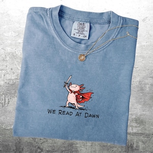 May include: Light blue Comfort Colors t-shirt with a graphic of an axolotl wearing a cape and holding a sword. The text "WE READ AT DAWN" is below the axolotl. A gold necklace with a pendant is draped on the shirt.