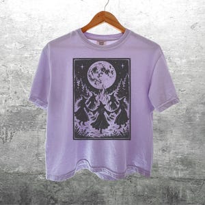 May include: A light purple short-sleeve t-shirt featuring a dark graphic print of four figures in pointed hats dancing in a circle around a bonfire beneath a large full moon and starry sky, with trees and foliage framing the scene.