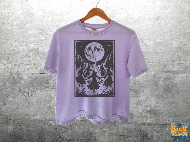 May include: A light purple short-sleeve t-shirt featuring a dark graphic print of four figures in pointed hats dancing in a circle around a bonfire beneath a large full moon and starry sky, with trees and foliage framing the scene.