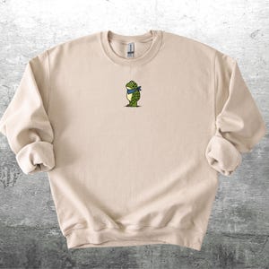 Puede incluir: Sudadera beige de cuello redondo con un peque&ntilde;o bordado de una rana con una bufanda azul. La rana es verde con manchas negras y est&aacute; de pie. La sudadera tiene pu&ntilde;os enrollados y cuello acanalado.