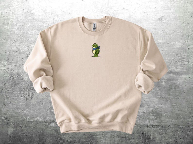 Puede incluir: Sudadera beige de cuello redondo con un peque&ntilde;o bordado de una rana con una bufanda azul. La rana es verde con manchas negras y est&aacute; de pie. La sudadera tiene pu&ntilde;os enrollados y cuello acanalado.